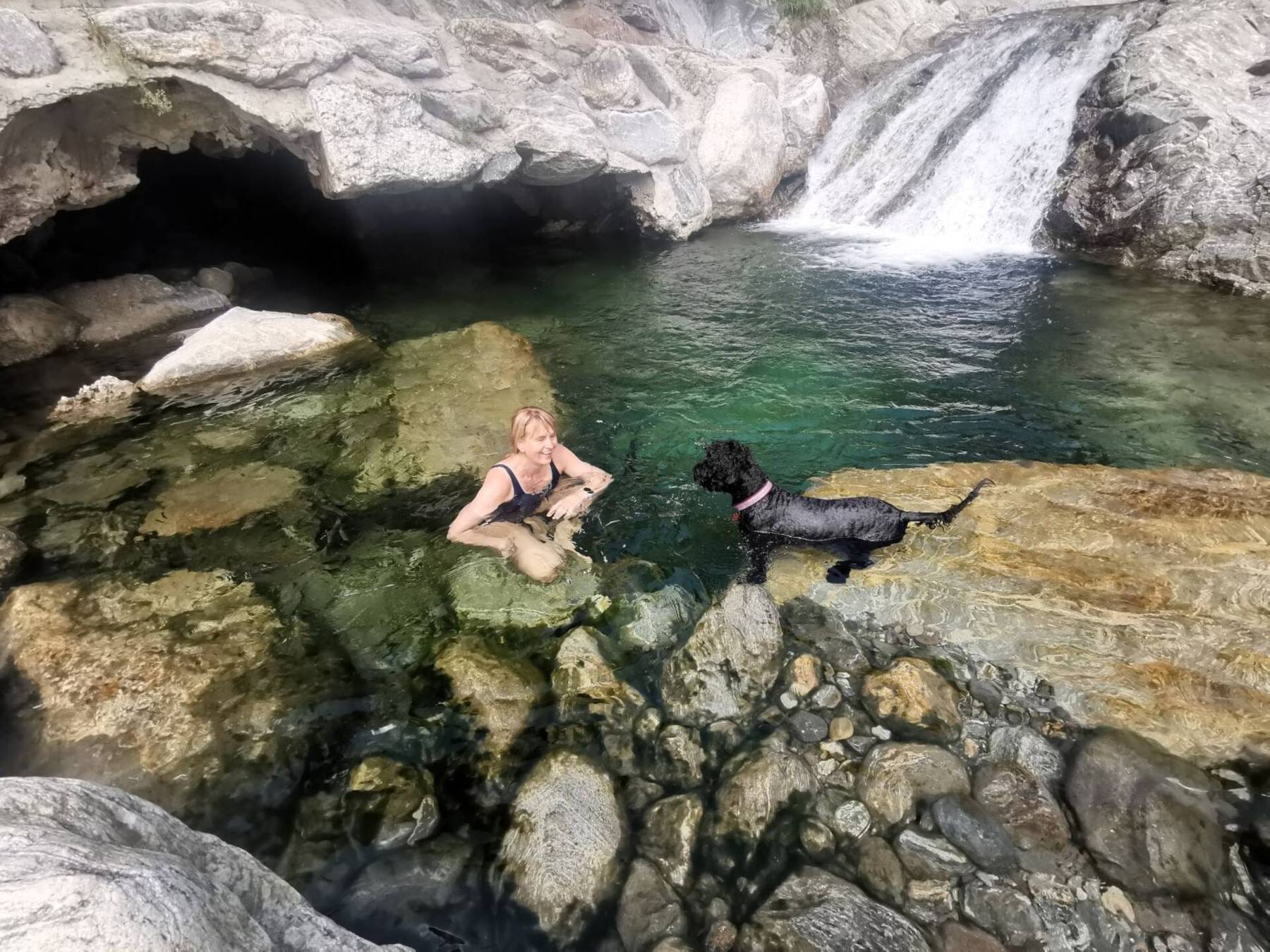 Badende og en svart hund ved en grønn fjellkulp under en liten foss